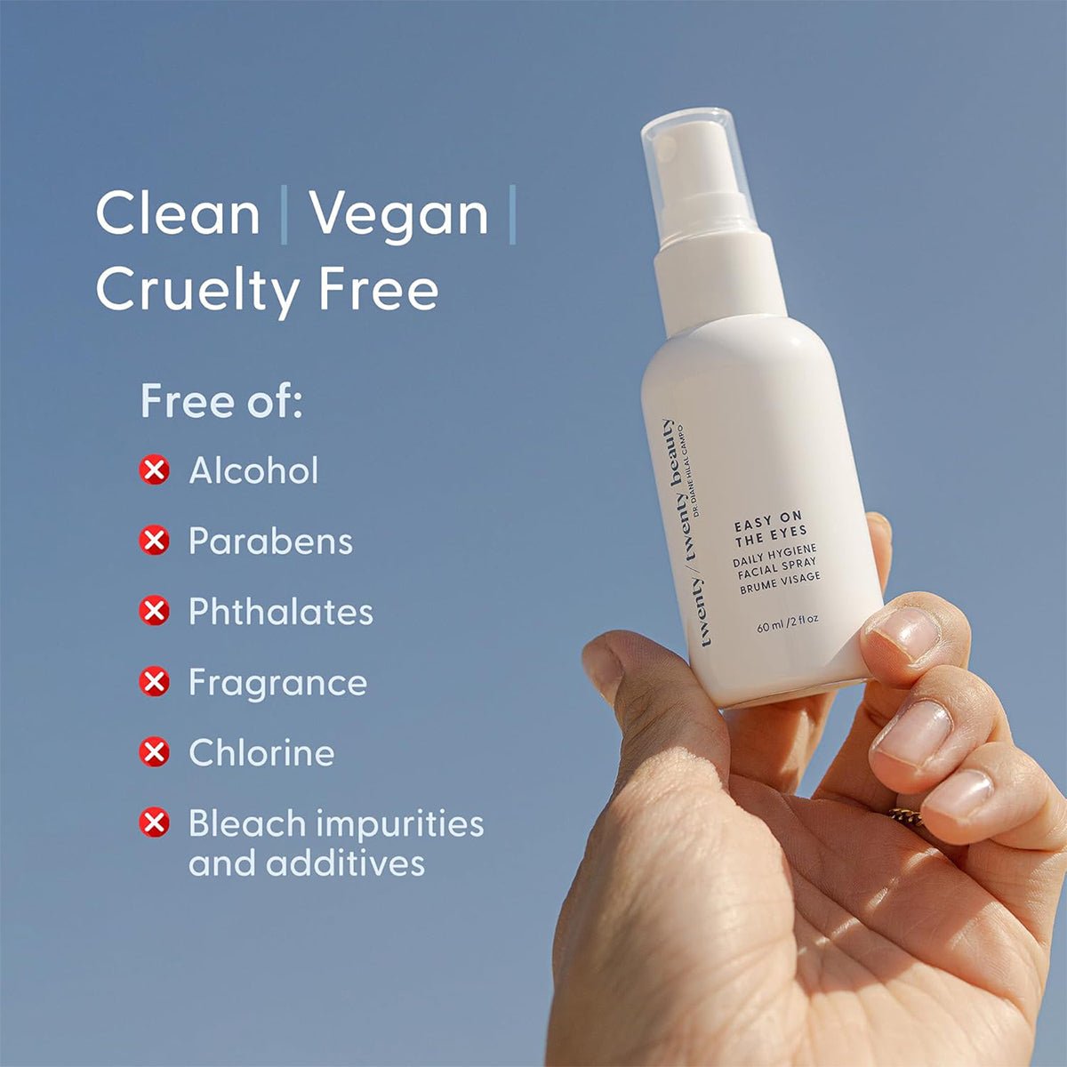 A hand holds a white spray bottle labeled Twenty Twenty Daily Hygiene Facial Spray. It is clean, vegan, cruelty-free, and free from alcohol, parabens, phthalates, fragrances, and chlorine. Developed by ophthalmologists with calming hypochlorous acid in a blue sky setting.