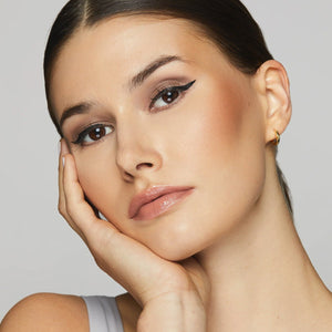 A person with long dark hair poses with their hand on their cheek, highlighting well-defined, ophthalmologist-tested makeup. Their eyes pop with winged eyeliner using Twenty Twenty Double Duty Precision Eyeliner (Pitch Black) 2.7mL, paired with neutral lipstick. A light top and small hoop earrings add sophistication.
