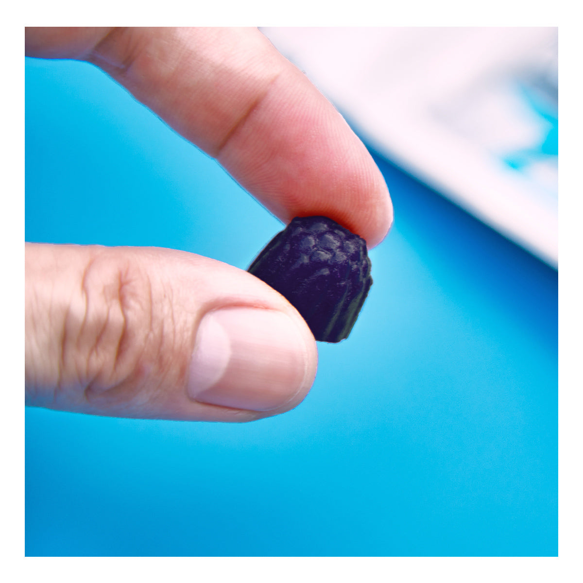 A hand holds a dark grape gummy against a bright blue background, reminiscent of the benefits FitEyez Eye Fitness Gummies offer for eye health.