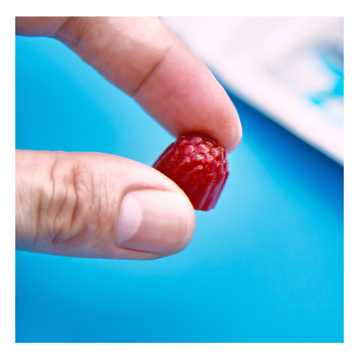 A hand holds a small, wild berry FitEyez gummy with a ridged surface against a bright blue background, highlighting the eye health nutrients in these Complete Multivitamin Eye Fitness Gummies by FitEyez.