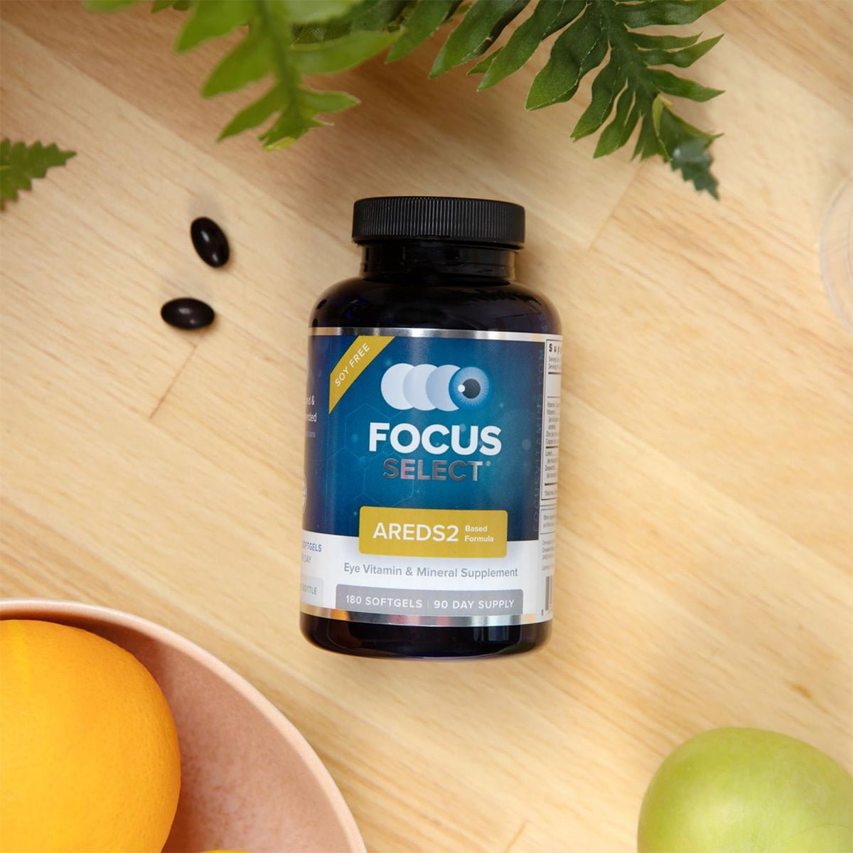 A bottle of Focus Select Soy Free Supplement by Focus Vitamins, featuring the AREDS2 formula for macular health, sits on a wooden surface with leaves and fruits. The black bottle with a yellow and blue label has two loose capsules nearby, showcasing this precision eye care product.