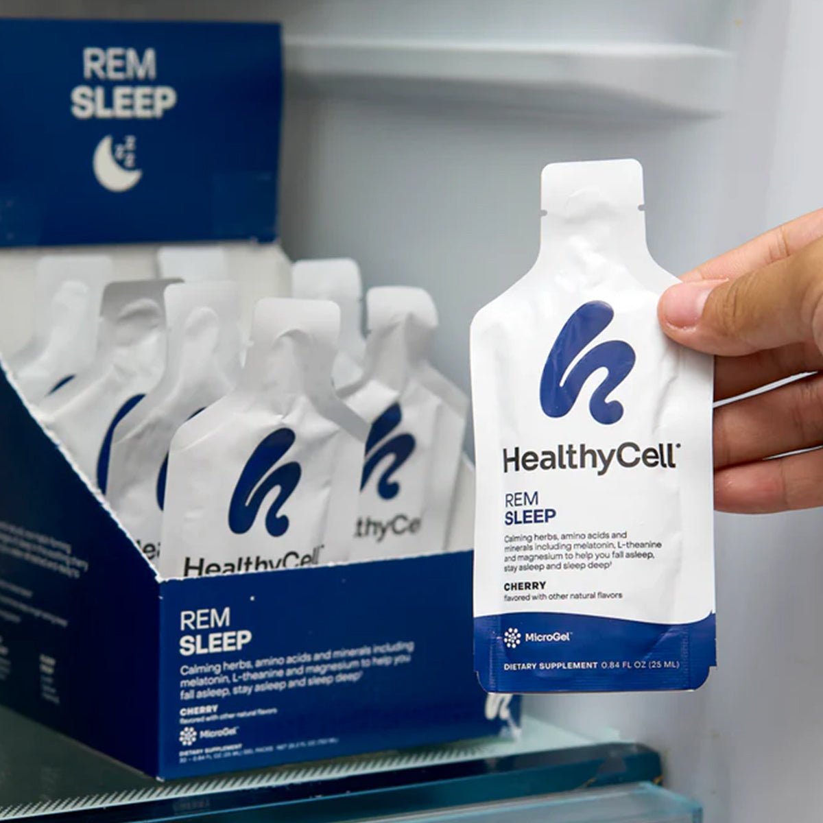 A hand reaches for a HealthyCell REM Sleep Supplement and Sleep Aid (30 Gel Packs) from a box. The white packs, featuring a blue logo, promise deep sleep support with calming herbs, amino acids, melatonin, and natural cherry flavor.