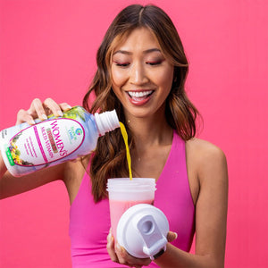 A woman in a pink tank top pours Tropical Oasis Womens Premium Multi-Vitamin Liquid Supplement (32oz.) into a shaker cup against a pink background, smiling and focused, embracing the anti-aging benefits of her daily routine.