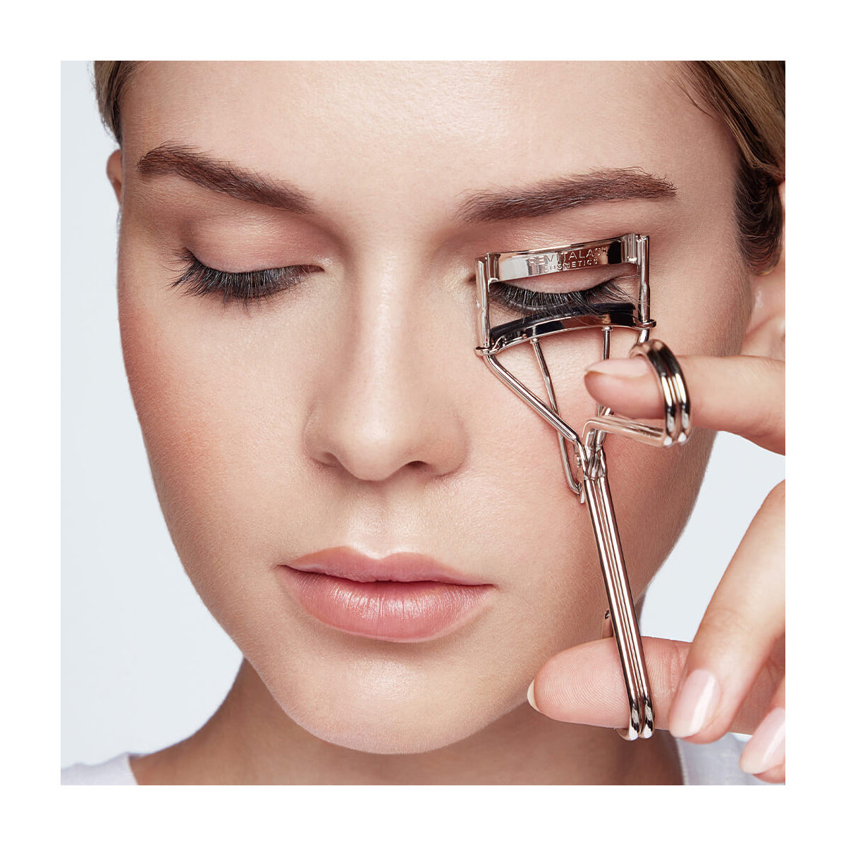 With a serene expression, a woman uses the RevitaLash Signature Eyelash Curler on her right eye. Her fair complexion and closed lips are highlighted against the white background, accentuating her graceful features and the sleek metal design of the RevitaLash tool.