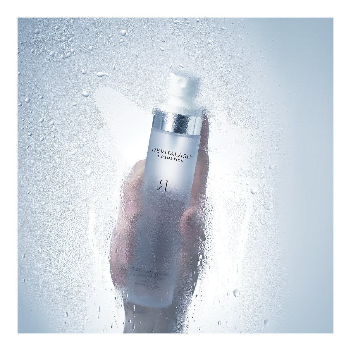 A hand holds a RevitaLash Micellar Water Lash Wash bottle against a foggy, wet glass. Partially covered in condensation, the 100mL hypoallergenic makeup remover from RevitaLash exudes freshness and cleanliness—ideal for sensitive eyes seeking gentle conditioning.