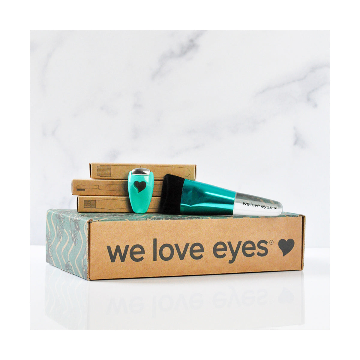 A stack of cardboard boxes with we love eyes features a teal bottle of We Love Eyes Complete Toolkit for Eyelashes and a small teal heart-shaped cup, all on pristine white marble, highlighting the importance of eyelid margin health.