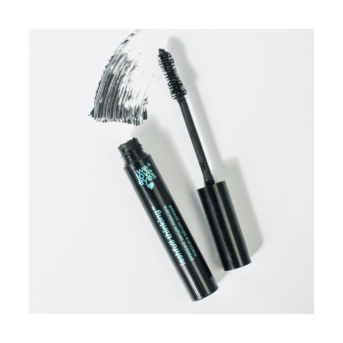 An open We Love Eyes Lashfull Thinking BLACK Pressed Serum Mascara tube, with its wand beside it, leaves a bold swipe on the white surface. The wand is coated with volumizing mascara enriched with botanical ingredients. The text on the tube reads We Love Eyes.