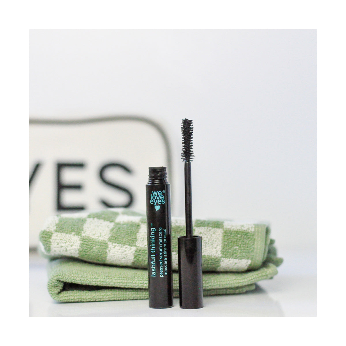 A tube of We Love Eyes Lashfull Thinking BLACK Pressed Serum Mascara with Widelash is placed in front of green checkered towels, with EYES visible on a sign, hinting at its lash growth formula. The mascara reads We Love Eyes.