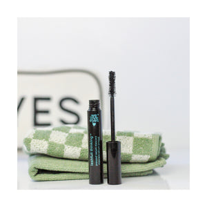 A tube of We Love Eyes Lashfull Thinking BLACK Pressed Serum Mascara with Widelash is placed in front of green checkered towels, with EYES visible on a sign, hinting at its lash growth formula. The mascara reads We Love Eyes.