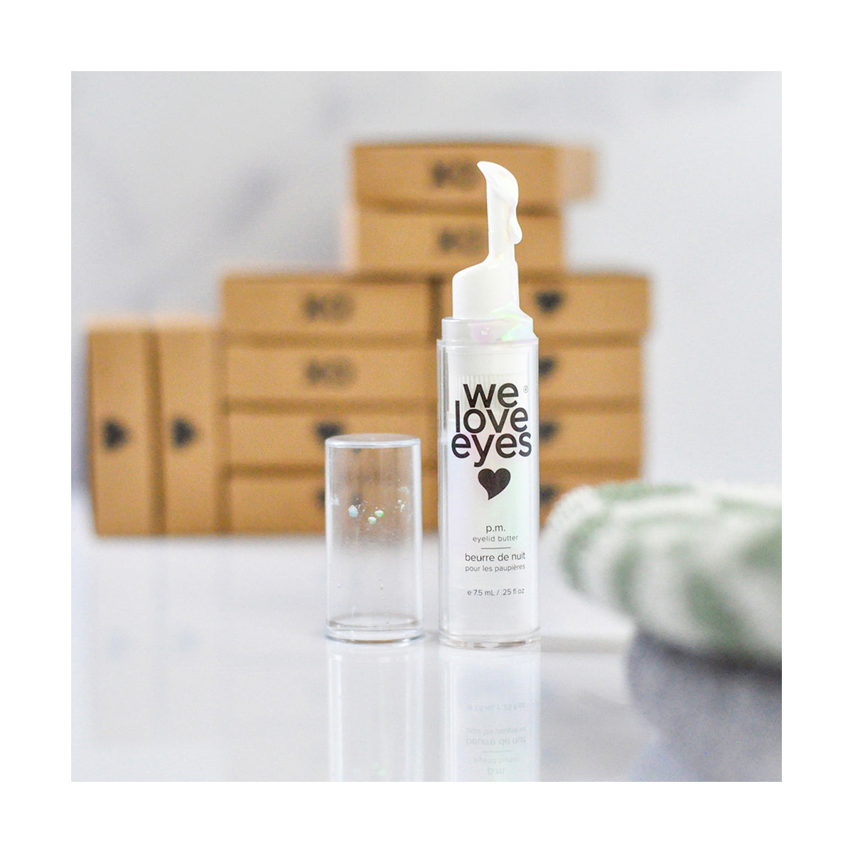 A bottle of We Love Eyes - P.M. Eyelid Butter - 7.5ml with a pump dispenser stands prominently, promoting meibomian gland health, next to its clear cap, while blurred behind are stacked brown boxes and green-white fabric touches.
