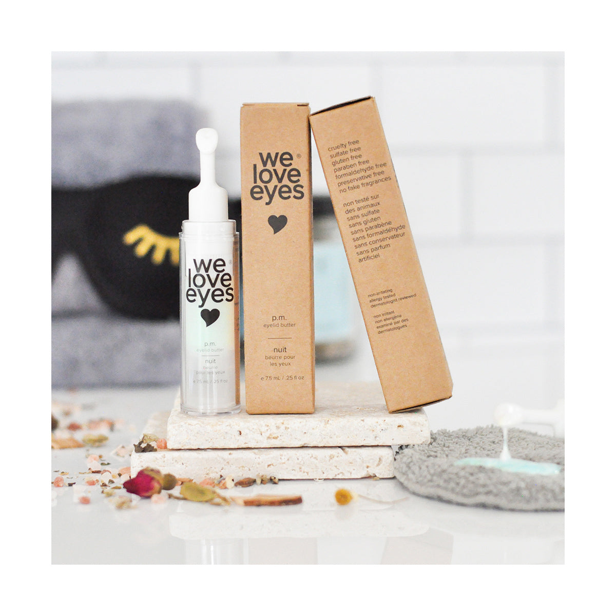 The We Love Eyes - P.M. Eyelid Butter (7.5ml) bottle sits on a bathroom counter, next to its brown box. Surrounded by scattered flowers and a gray washcloth, an eye mask is blurred in the background, accentuating the products oil-based support for meibomian gland health.