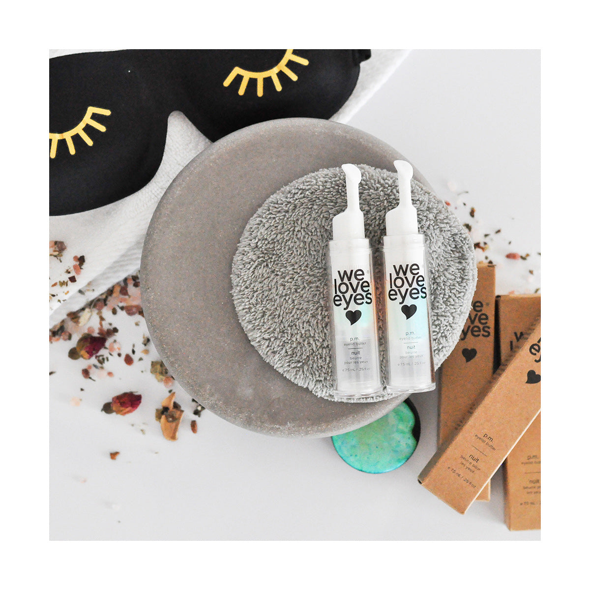 Two bottles of We Love Eyes - P.M. Eyelid Butter sit on a gray pad, surrounded by brown boxes, a black eye mask with eyelash designs, and dried petals, highlighting the focus on meibomian gland health and reducing transepidermal water loss.