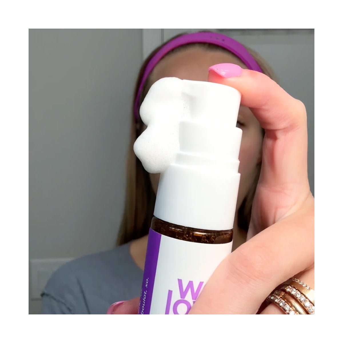 A person with manicured nails and a pink headband presses down on a We Love Eyes SuperGLA eyelid & eyelash foaming cleanser bottle, dispensing non-toxic white foam. The bottles white and purple label highlights its gentle formula.