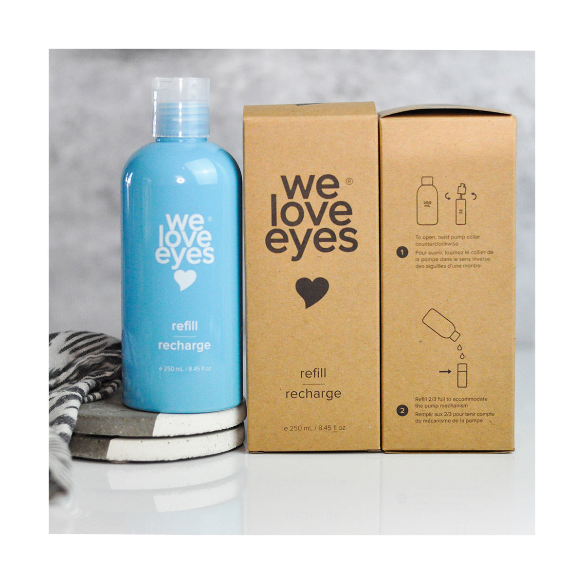 A blue 250mL We Love Eyes Tea Tree Eyelid Foaming Cleanser Refill bottle, made from Post-Consumer Recycled Plastic, labeled refill recharge, is beside two brown boxes with branding, instructions, and a heart symbol. A striped cloth and round pads lie nearby.