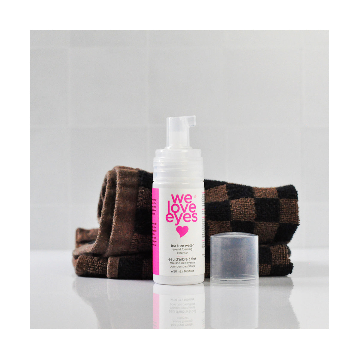 A bottle of We Love Eyes Tea Tree Water Eyelid Foaming Cleanser is placed on a reflective surface in front of a folded brown checkered towel against a tiled wall. The pH-balanced formula, known for gentle eye care, has its cap resting beside it.