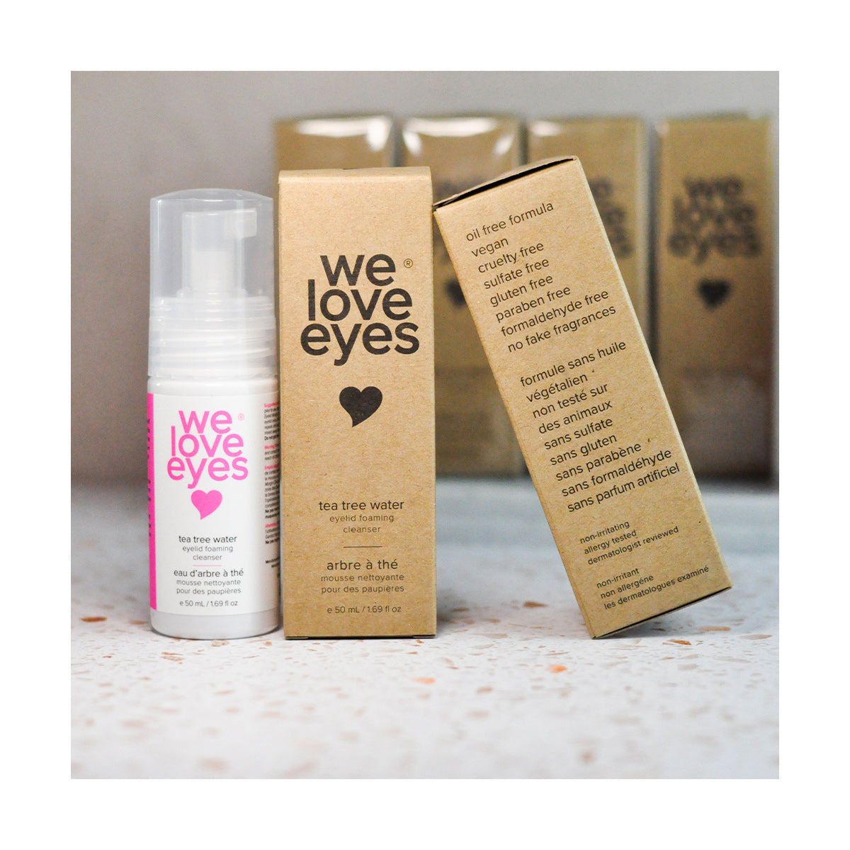 A bottle and two boxes of We Love Eyes Tea Tree Water Eyelid Foaming Cleanser rest on a speckled surface. The white bottle has pink text, and the brown boxes highlight ingredients and benefits. This pH-balanced solution offers gentle eye care.