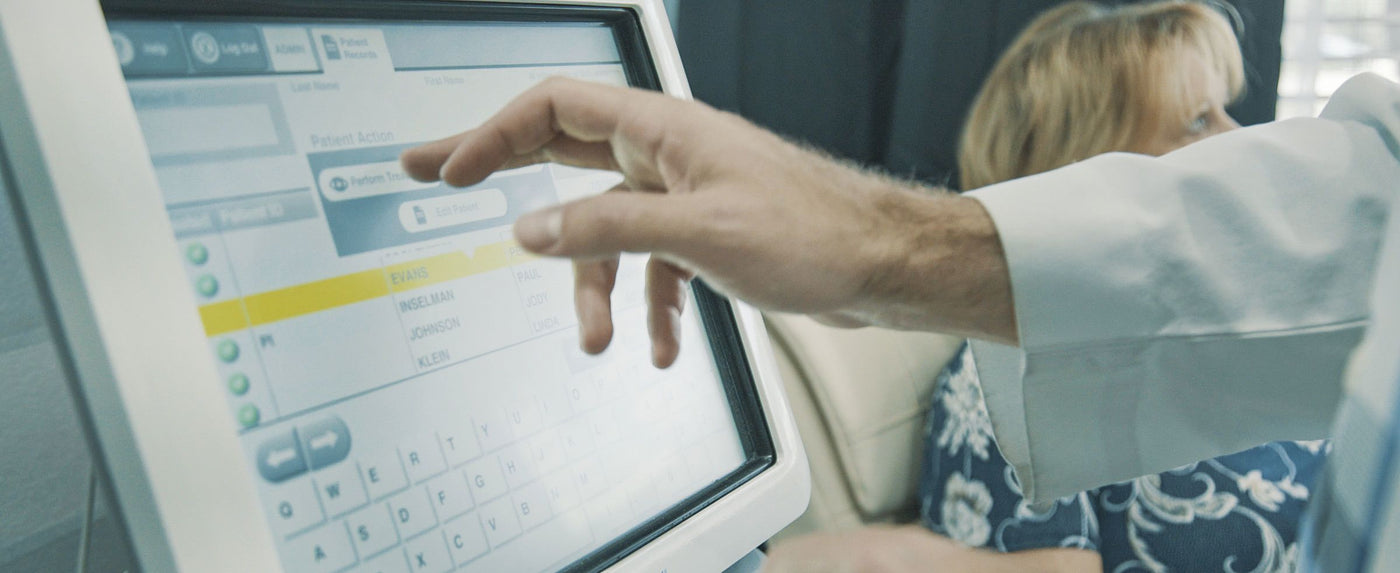 A person wearing a white lab coat is using a touchscreen device, selecting options from a digital menu. Another person is seated nearby, partially visible, dressed in a patterned outfit.