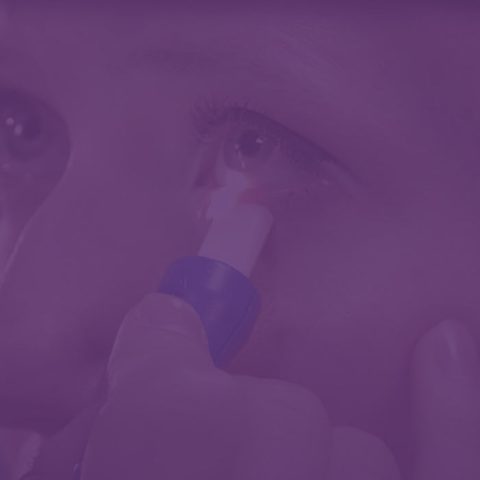 Close-up of a person applying a small cylindrical stick to their lower eyelid. The image has a purple tint, with focus on the eye and the applicator.