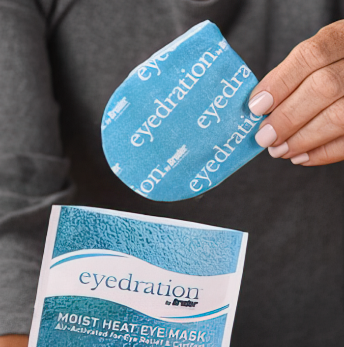 A person holds a blue eye mask packet labeled eyedration. They are placing or removing an eye mask from the open packet. The packaging mentions Moist Heat Eye Mask along with other details.