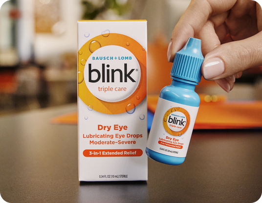 A hand holds a small blue bottle labeled Blink Triple Care next to its box. The box reads Bausch + Lomb Blink Triple Care, Dry Eye Lubricating Eye Drops, Moderate-Severe, 3-in-1 Extended Relief.