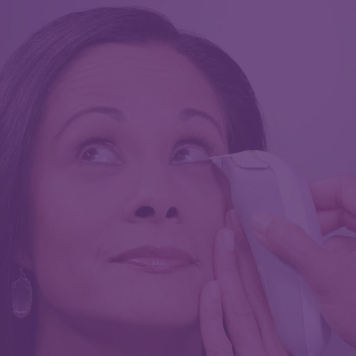 A person with long hair is having a procedure done near their eye using a handheld device. Another persons hands are holding the device gently against the persons face. The image has a purple tint.
